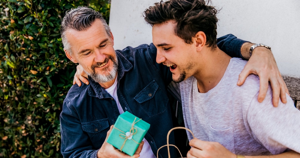 A father and son laughing and bonding over a teal gift box outside.