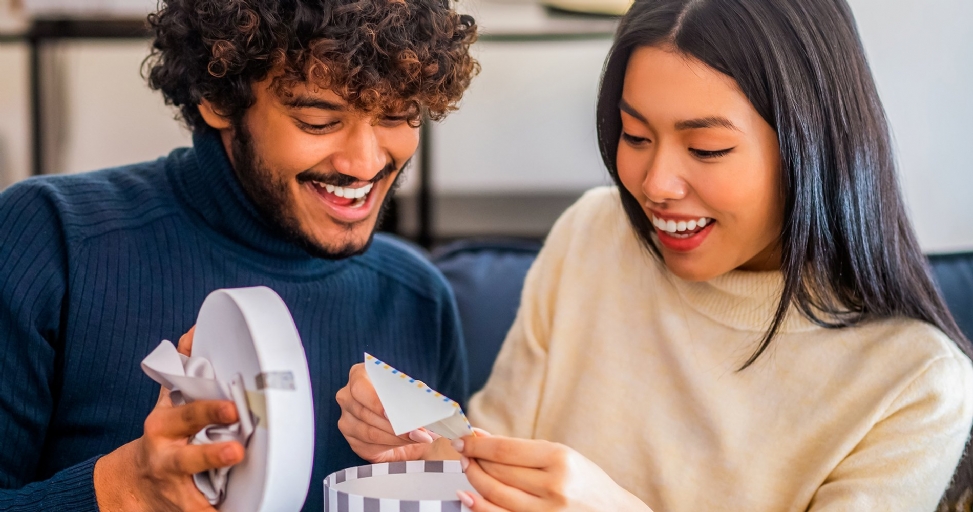 A happy couple smiles while opening a round gift box, revealing a small card inside.