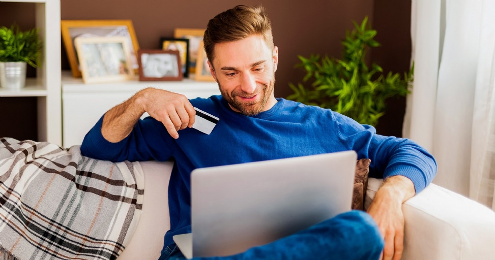Smiling man on a couch uses a laptop and holds a credit card for online shopping.