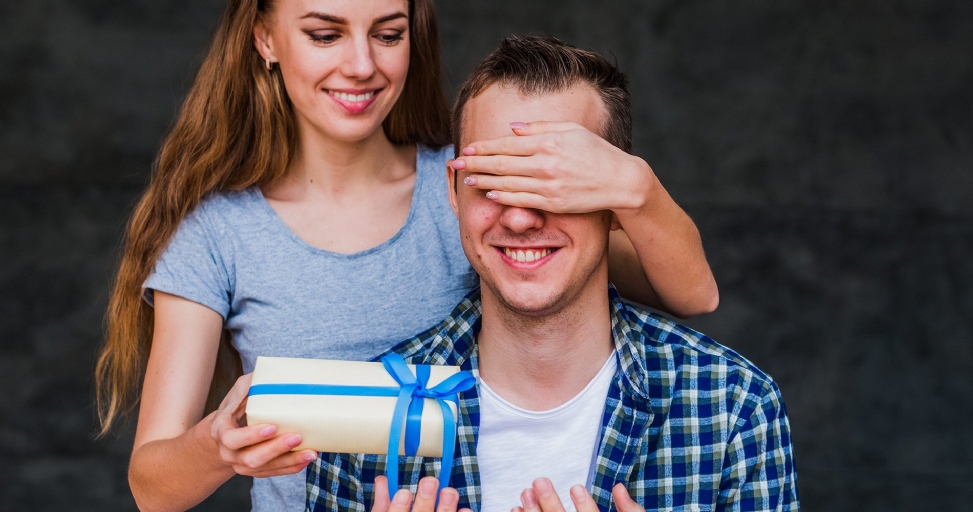 A smiling woman surprises a man by covering his eyes and presenting a wrapped gift.