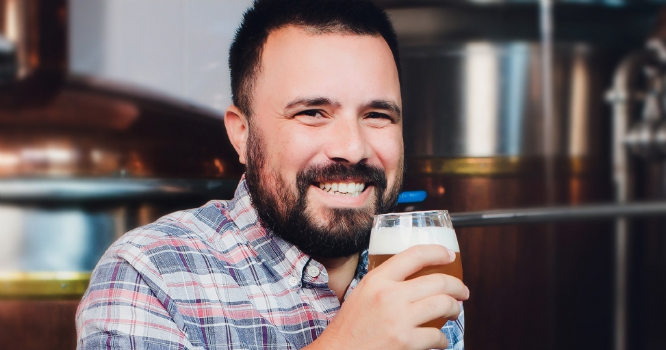 A happy bearded man in a plaid shirt smiles while holding a glass of beer in a brewery.