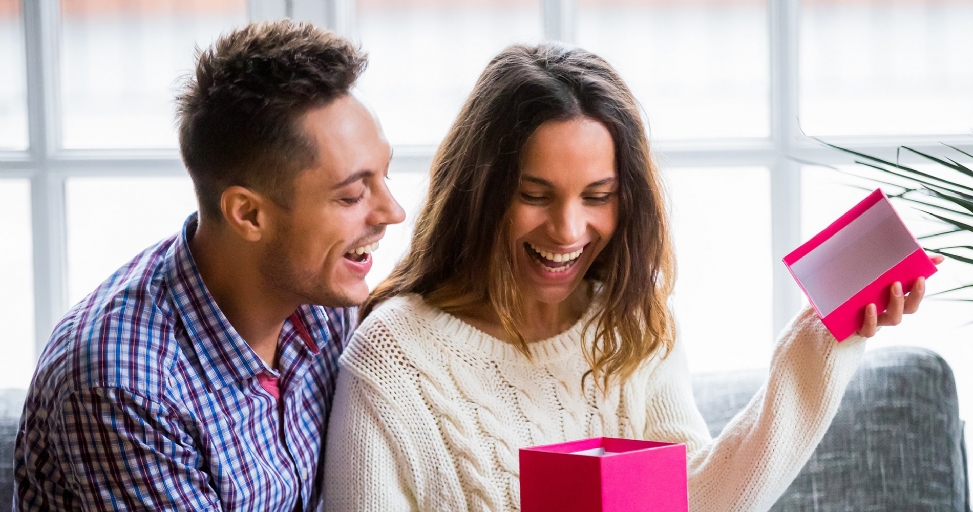 A joyous couple shares a laugh as the woman excitedly opens a pink gift box.