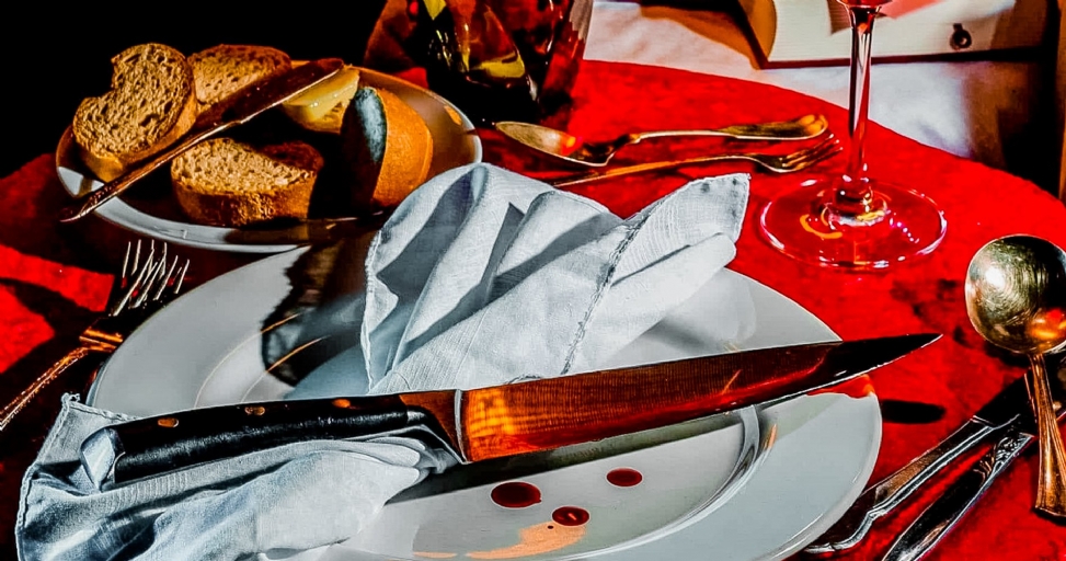 A dinner table setting with a red tablecloth, white plates, and a large knife with blood drops.