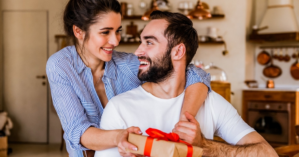 Smiling couple in a home, woman hugging man who holds a gift with red ribbon.