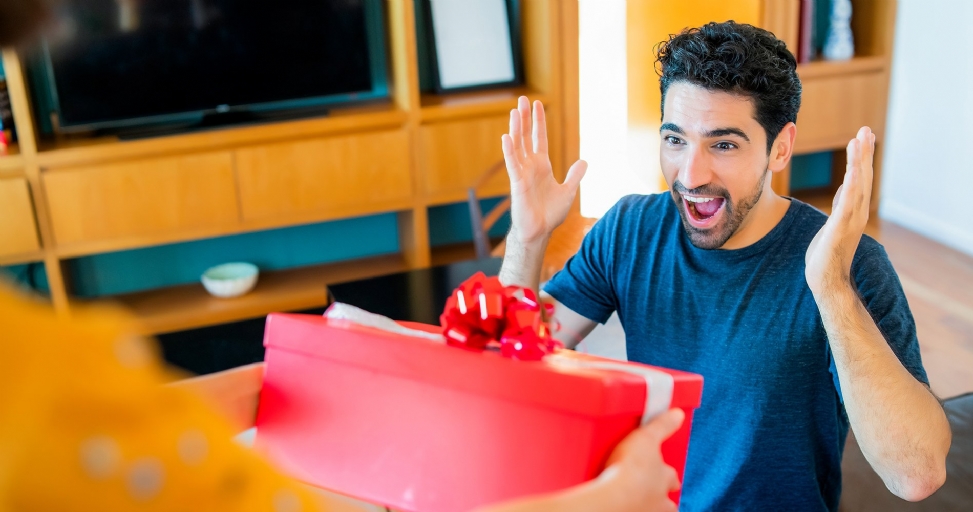 Excited man with raised hands receiving a red gift box from another person in a living room.
