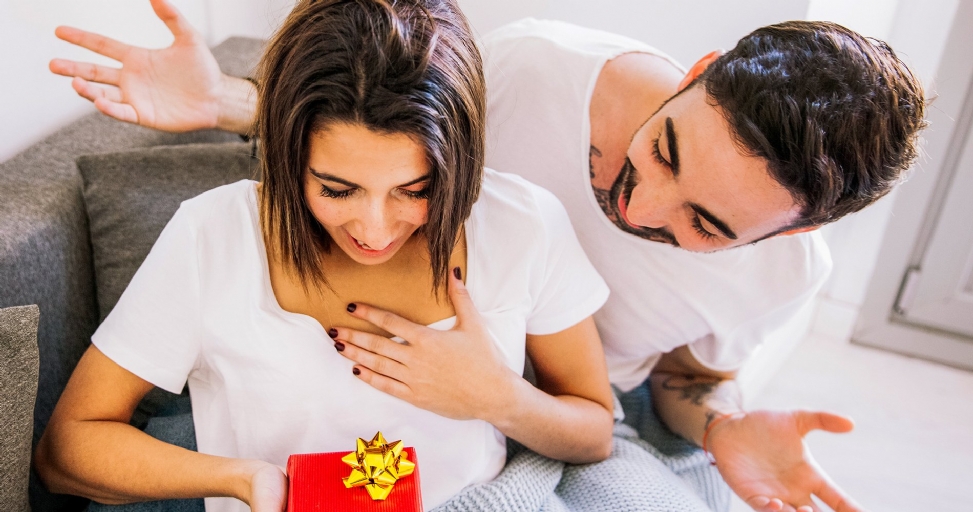 A happy woman receives a red gift box with a golden bow from a smiling man.