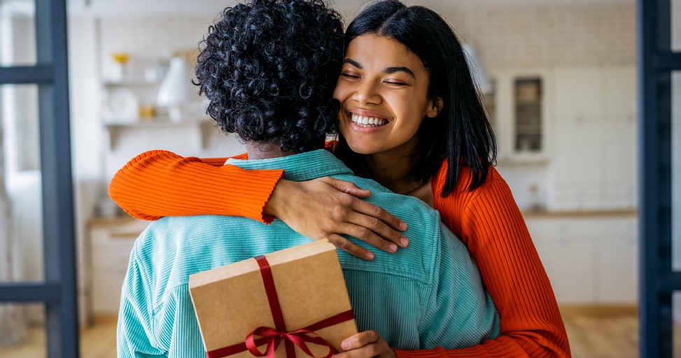 A joyful woman in an orange sweater hugging a person holding a thoughtful gift box.