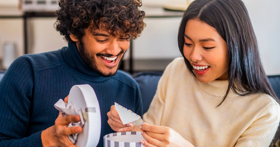 A happy couple opening gifts, smiling and looking at a small card inside a gift box.