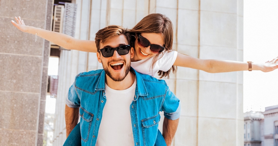A cheerful man giving a piggyback ride to a happy woman with outstretched arms, both smiling.