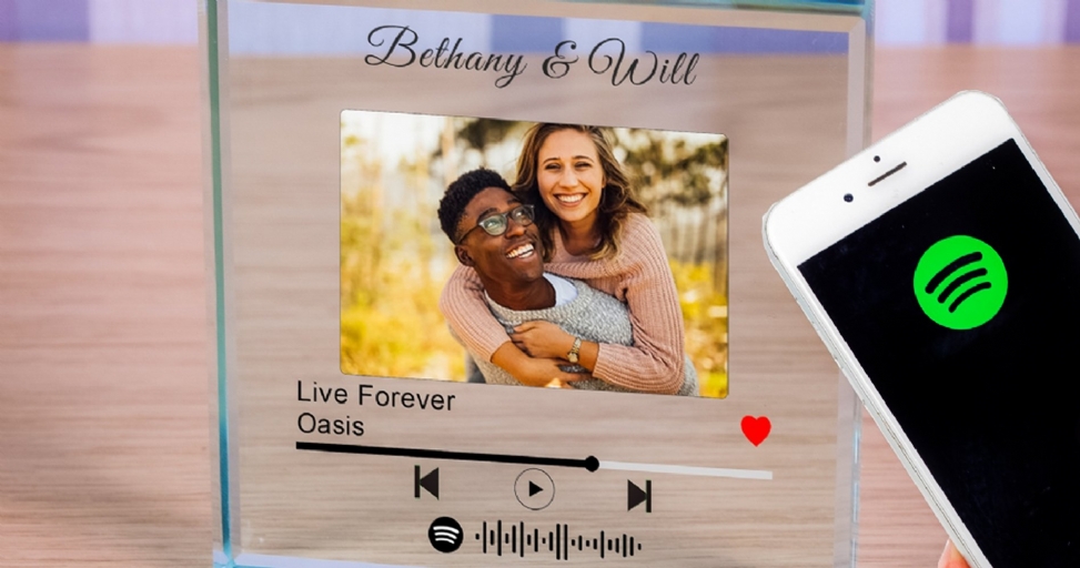 A personalized Spotify glass plaque featuring a smiling couple, the song 'Live Forever' by Oasis, and a Spotify code, next to a phone displaying the Spotify logo.