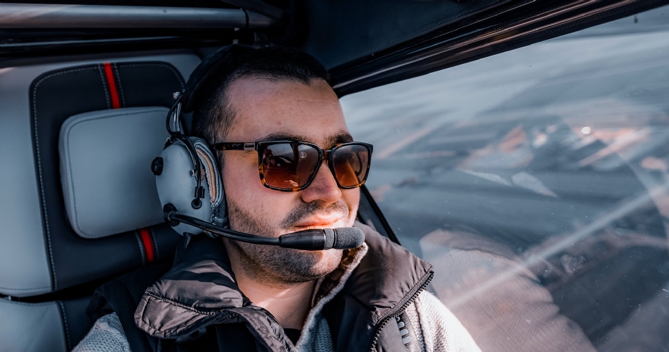Close-up of a man wearing sunglasses and a pilot headset in a cockpit, looking out the window.