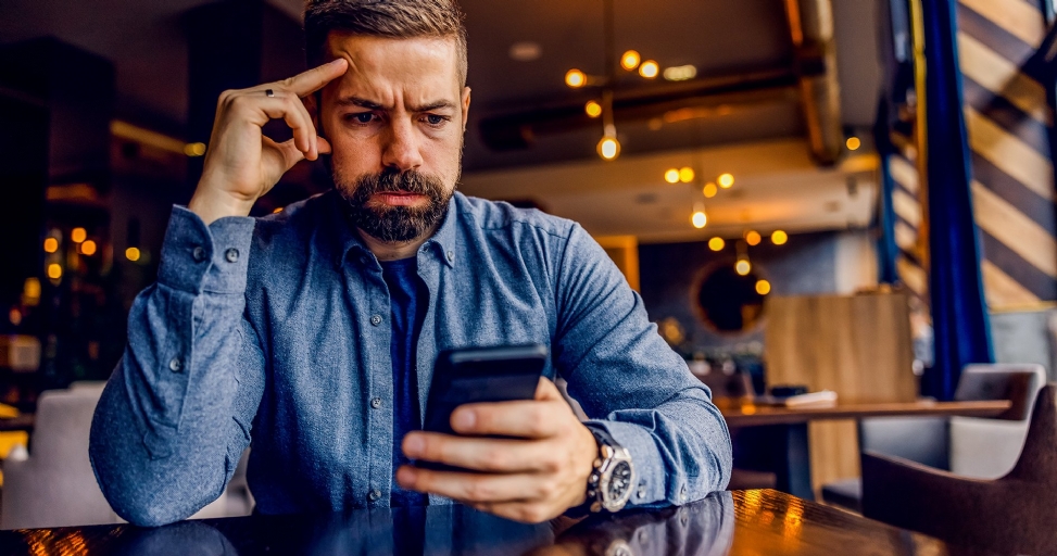 A bearded man looks intently at his phone with a concerned expression, hand to his head.
