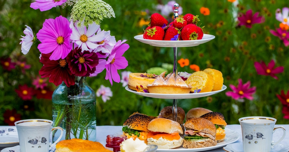 An outdoor afternoon tea spread with pastries, sandwiches, strawberries, flowers, and teacups in a garden setting.