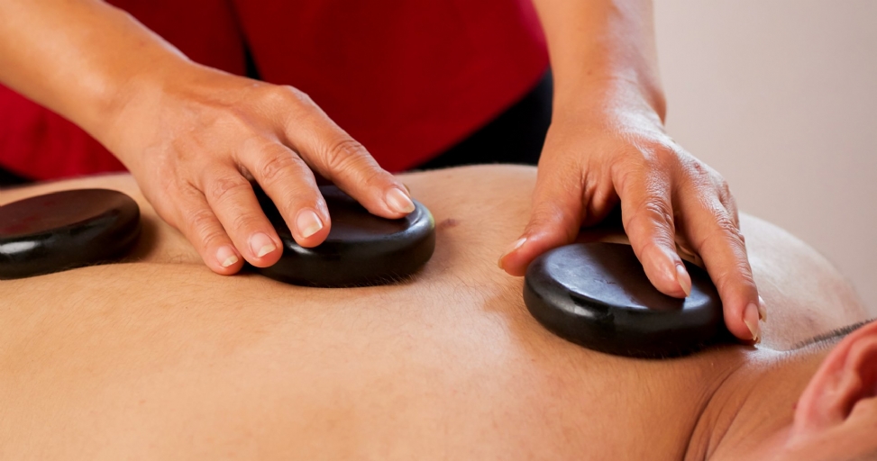 A person receives a relaxing hot stone massage, with a masseuse's hands placing warm, dark stones on their back.