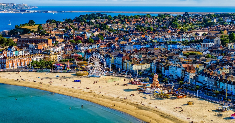 Aerial view of a vibrant coastal town with a sandy beach, Ferris wheel, amusement rides, and colorful buildings by the sea.