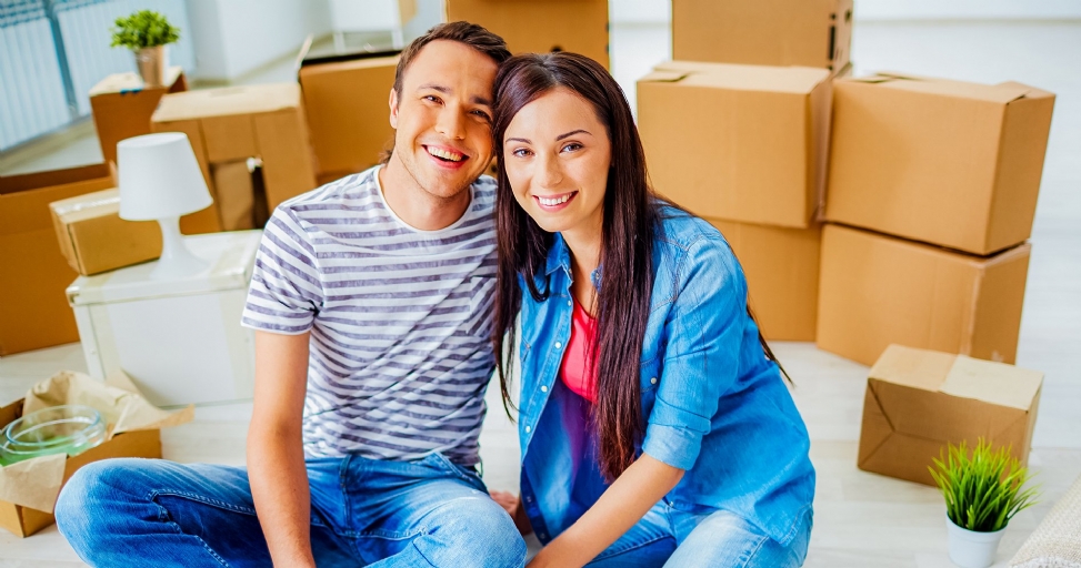 A happy smiling couple sits on the floor surrounded by numerous cardboard moving boxes.