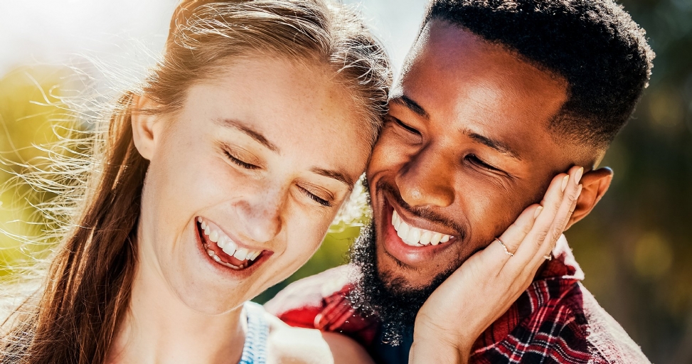 A joyful couple, eyes closed and laughing, sharing a tender moment in bright sunlight.