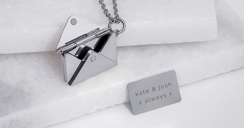 A silver envelope locket, open to reveal its interior, next to a personalized 'Kate & Josh' plaque.