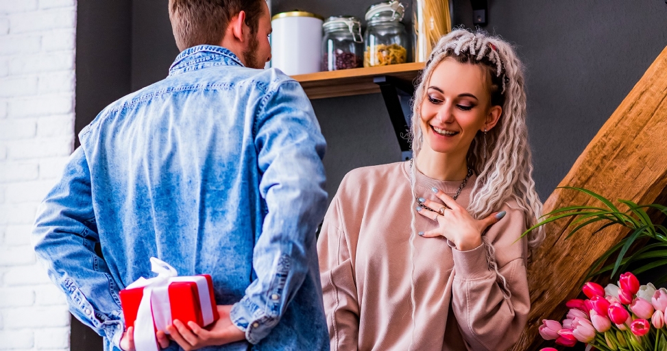 A man surprises a smiling woman with a hidden red gift, holding it behind his back.