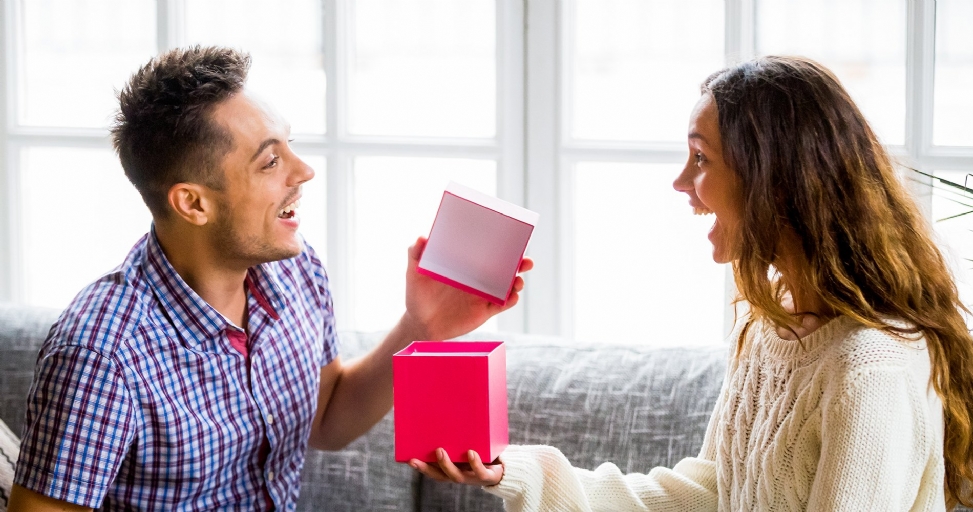 A man surprises a woman with an open gift box, both smiling with excited expressions.