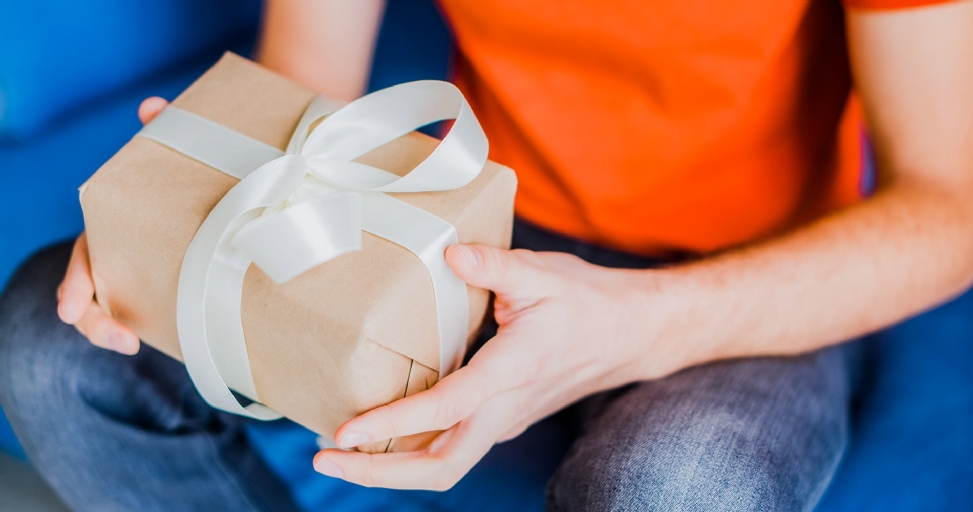 A person in an orange shirt and blue jeans holds a brown paper gift with a white ribbon.