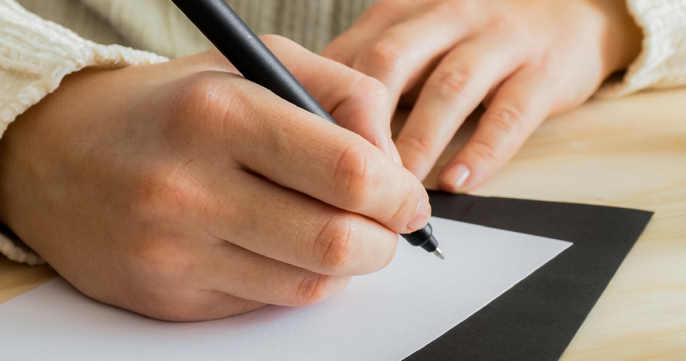 Close-up of a person's hands writing on white paper with a black pen.