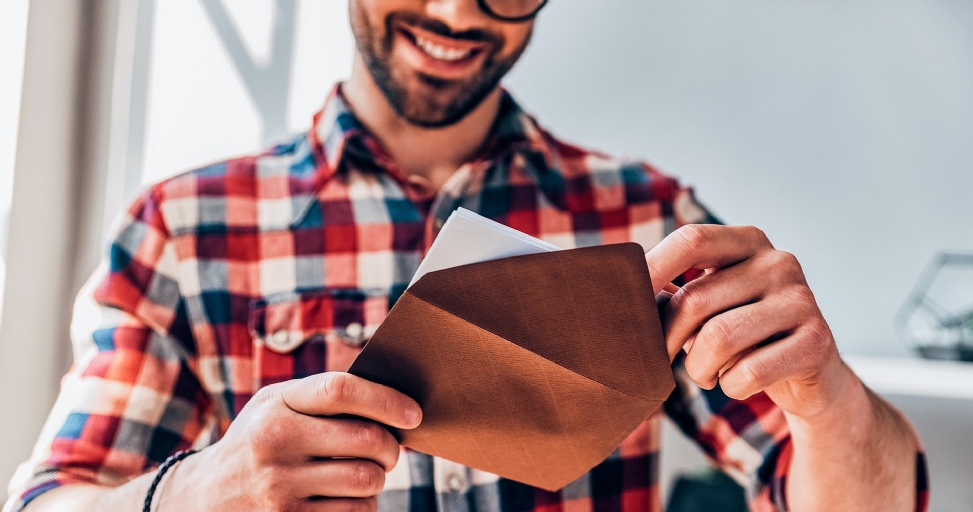 A smiling man in a plaid shirt holds an open brown envelope with white cards inside.