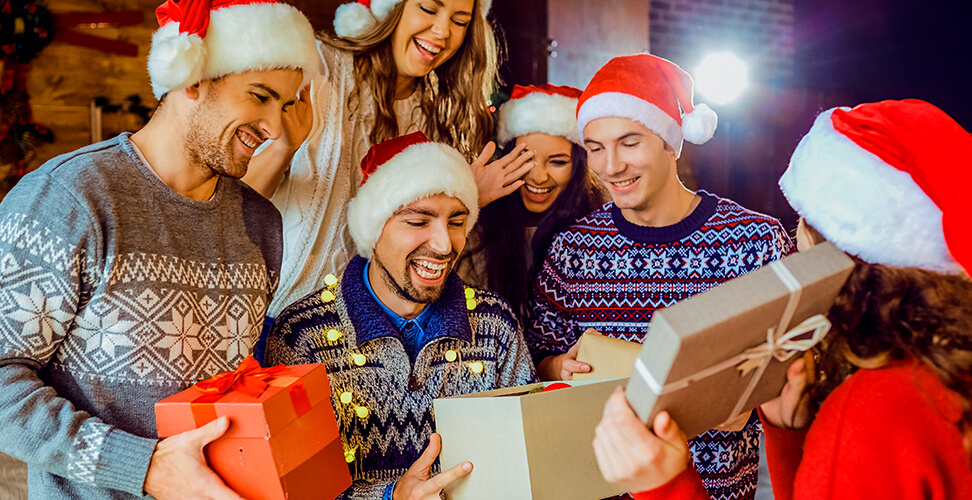 Group of friends in Christmas jumpers laughing and opening White Elephant gifts at holiday party